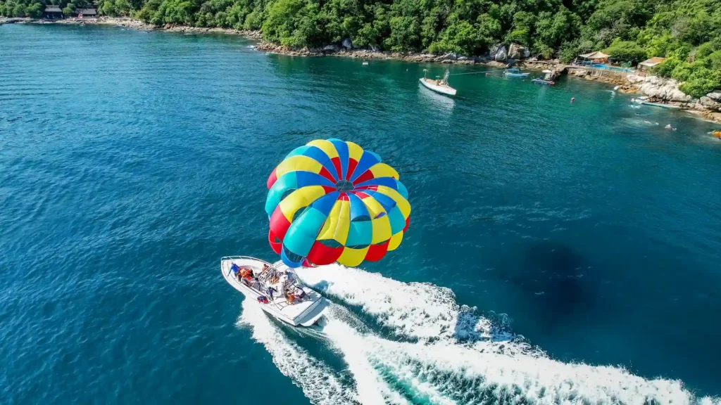 Vista aérea de una lancha tirando un paracaídas multicolor con personas practicando paravelismo sobre el mar azul en Bahía de Banderas.
