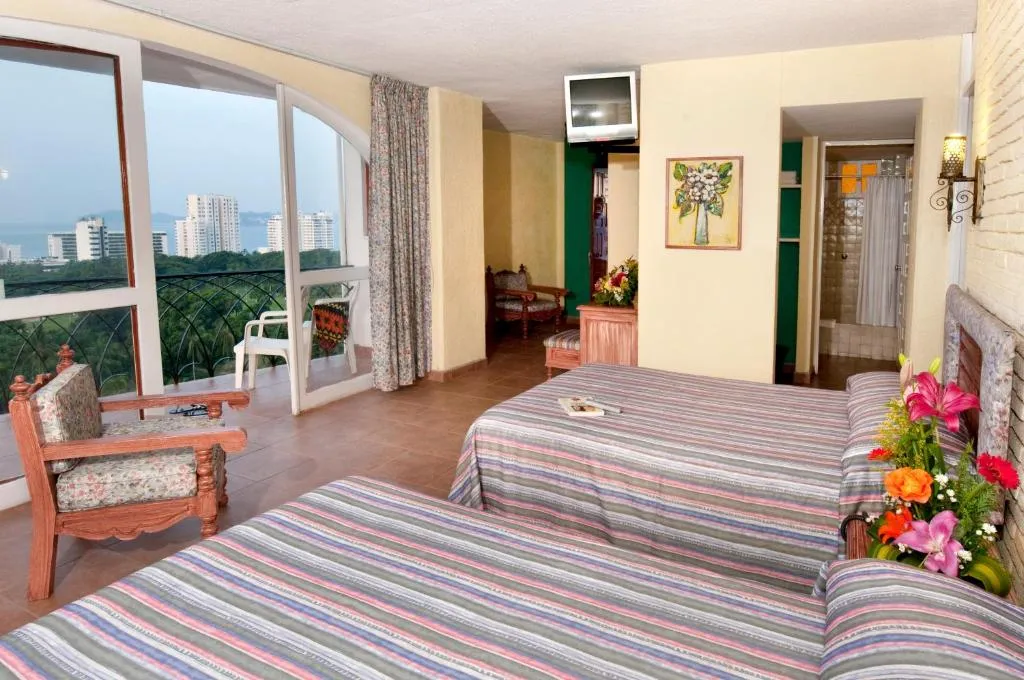 Habitación de hotel con dos camas dobles, balcón con vista a la ciudad y edificios al fondo, decoración sencilla con muebles de madera, flores frescas sobre la cama y una televisión montada en la pared.