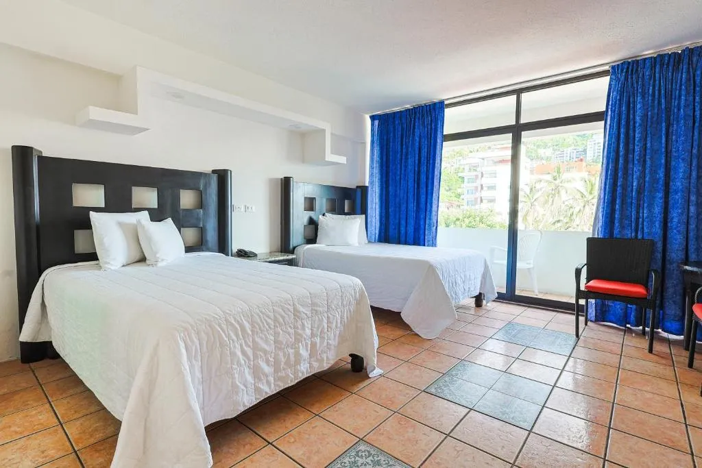 Habitación de hotel con dos camas individuales, piso de baldosas, cortinas azules, balcón con sillas y vista a palmeras y edificios al fondo.