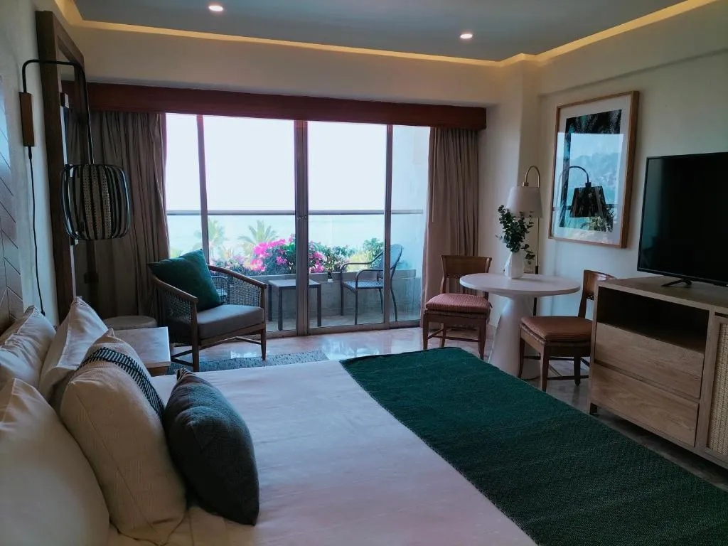 Suite de hotel elegante con cama matrimonial, decoración moderna en tonos neutros, balcón con flores y vista al océano, mesa con sillas y televisión de pantalla plana.