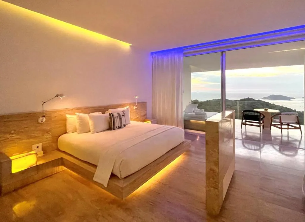 Suite de hotel moderna con cama matrimonial, iluminación cálida, balcón con ventanales de vidrio y vista panorámica al océano y colinas al atardecer.